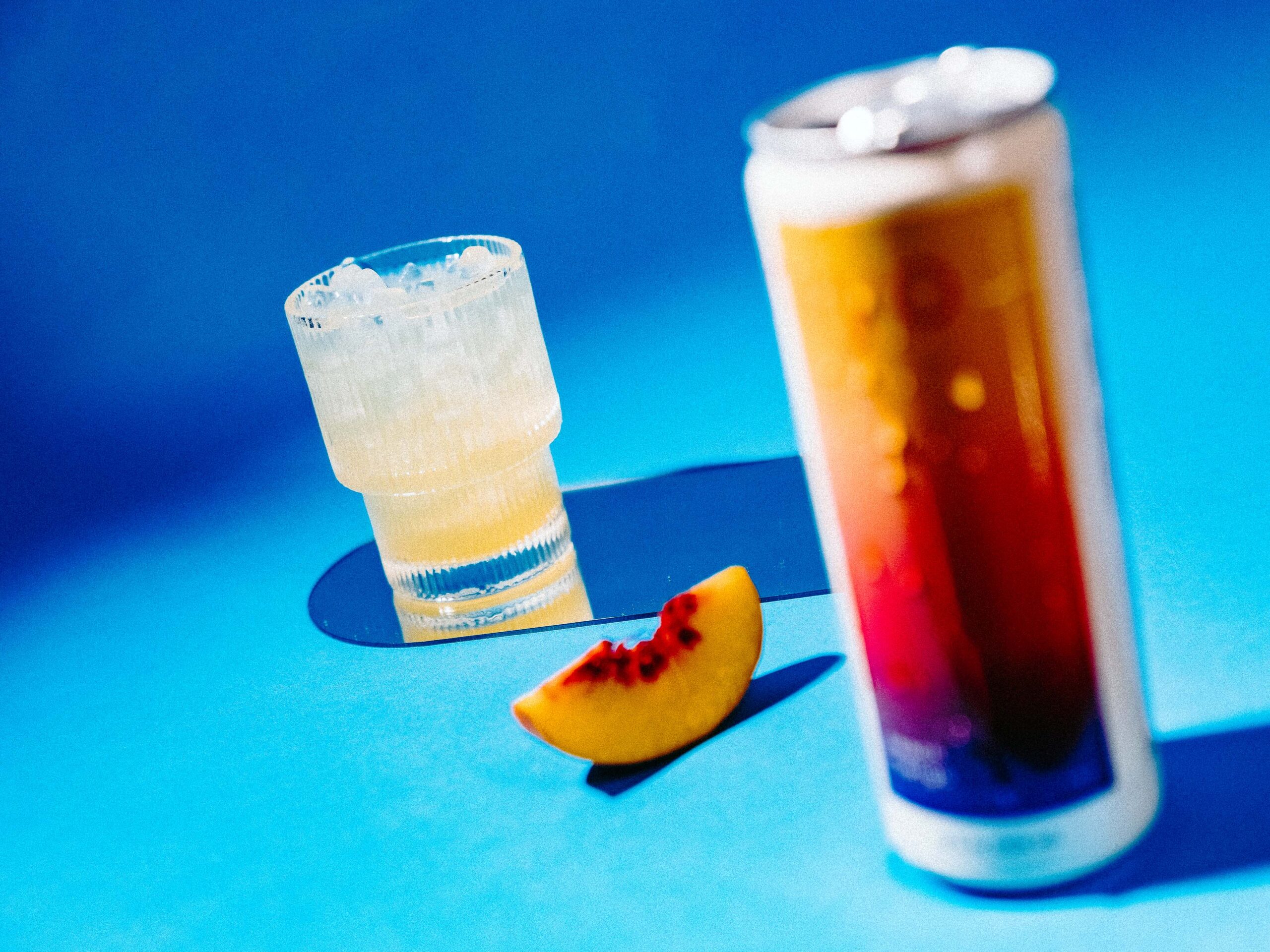 A can of Peach Mango Hiyo sits closest to the camera, with a slice of peach and iced glass of Hiyo sitting behind it.