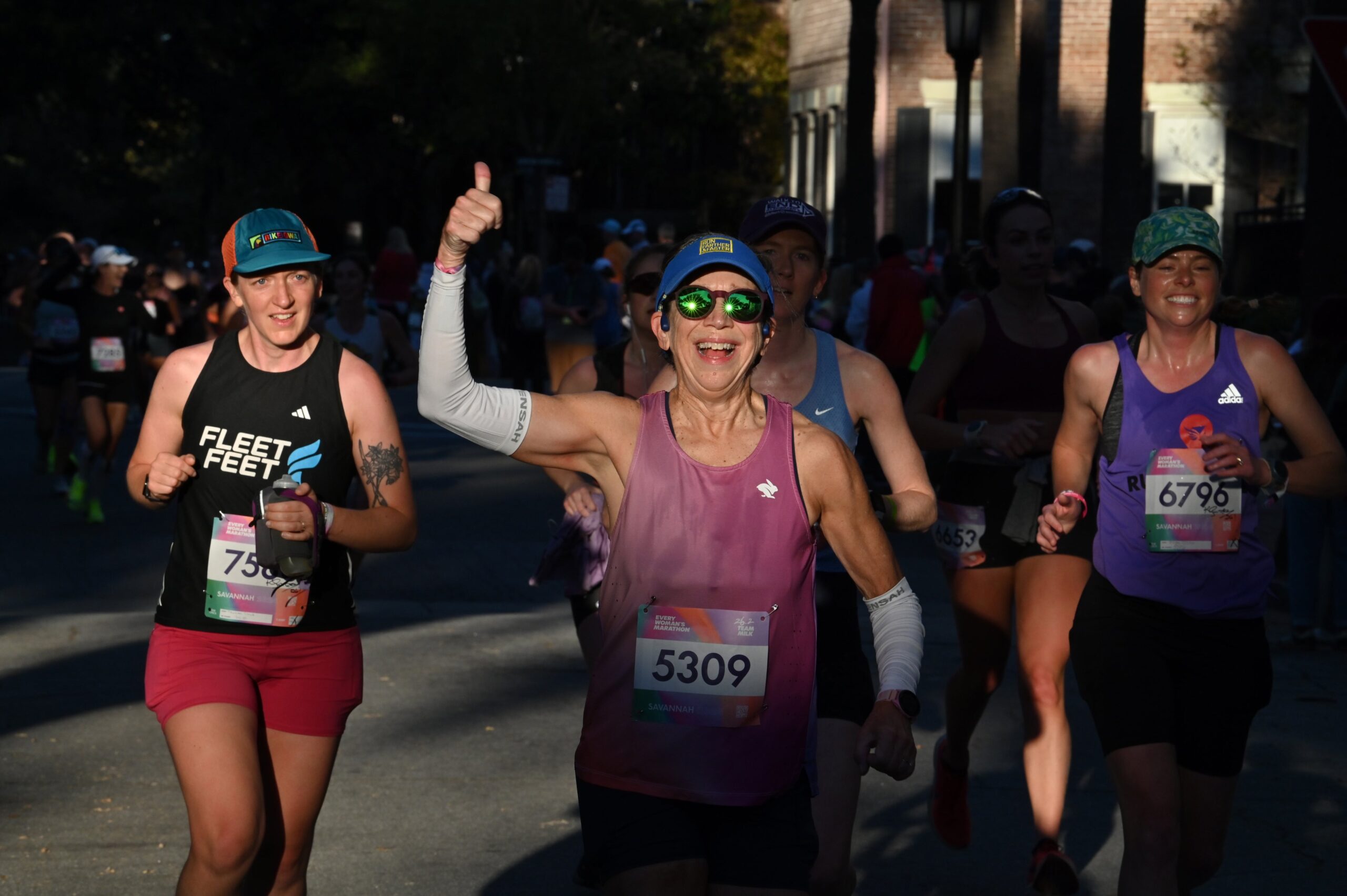 Monica, founder of rabbit, offers a thumbs up to the camera while running in an all-women's marathon alongside a group of women.