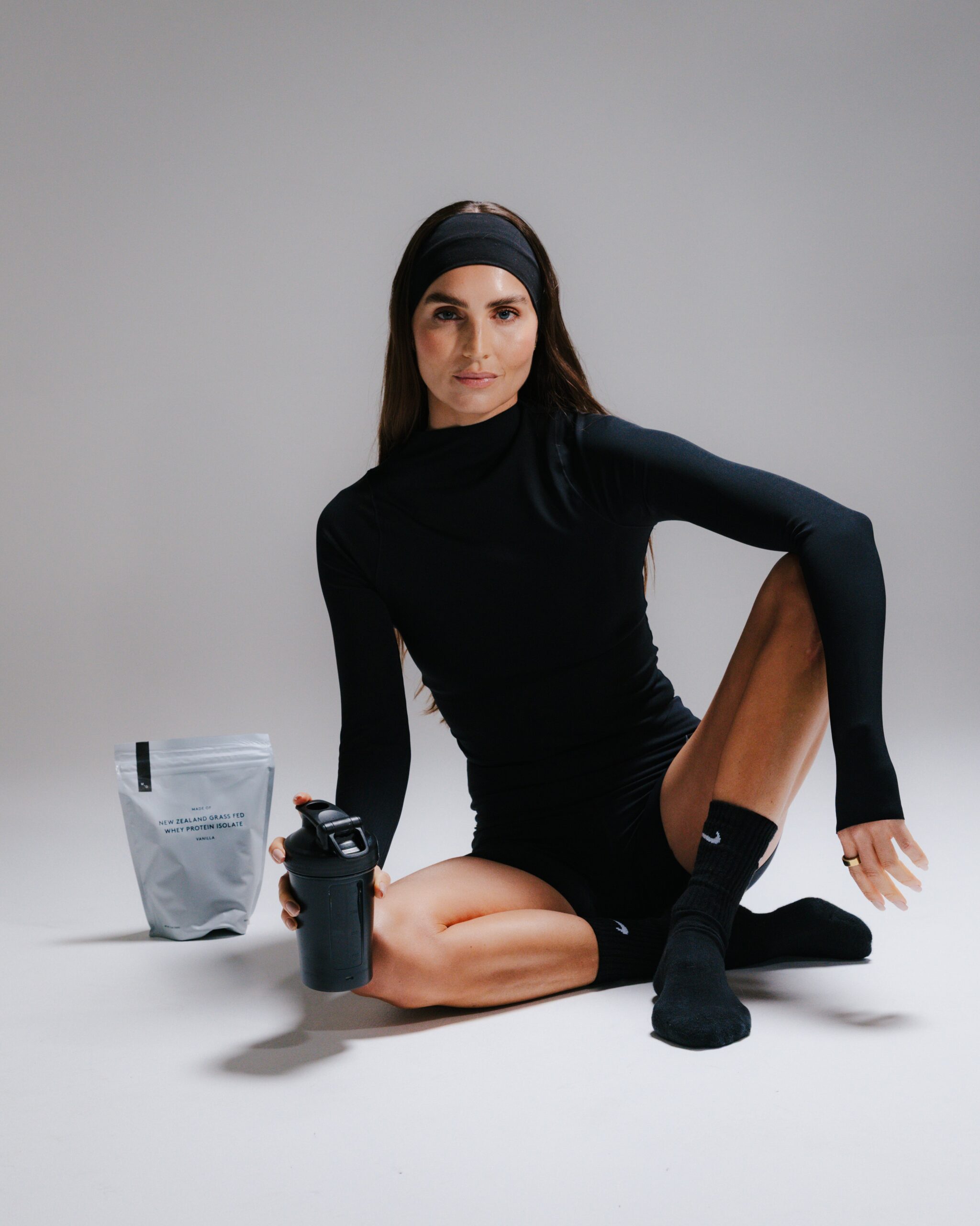 Kirsty Godso sitting on the floor holding a shaker bottle next to a bag of VANILLA WHEY PROTEIN ISOLATE.