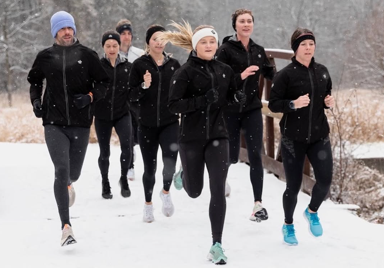 A group of runners brave the elements as they run across a snowy trail, wearing black rabbit running jackets, headbands, and gear designed for winter running.