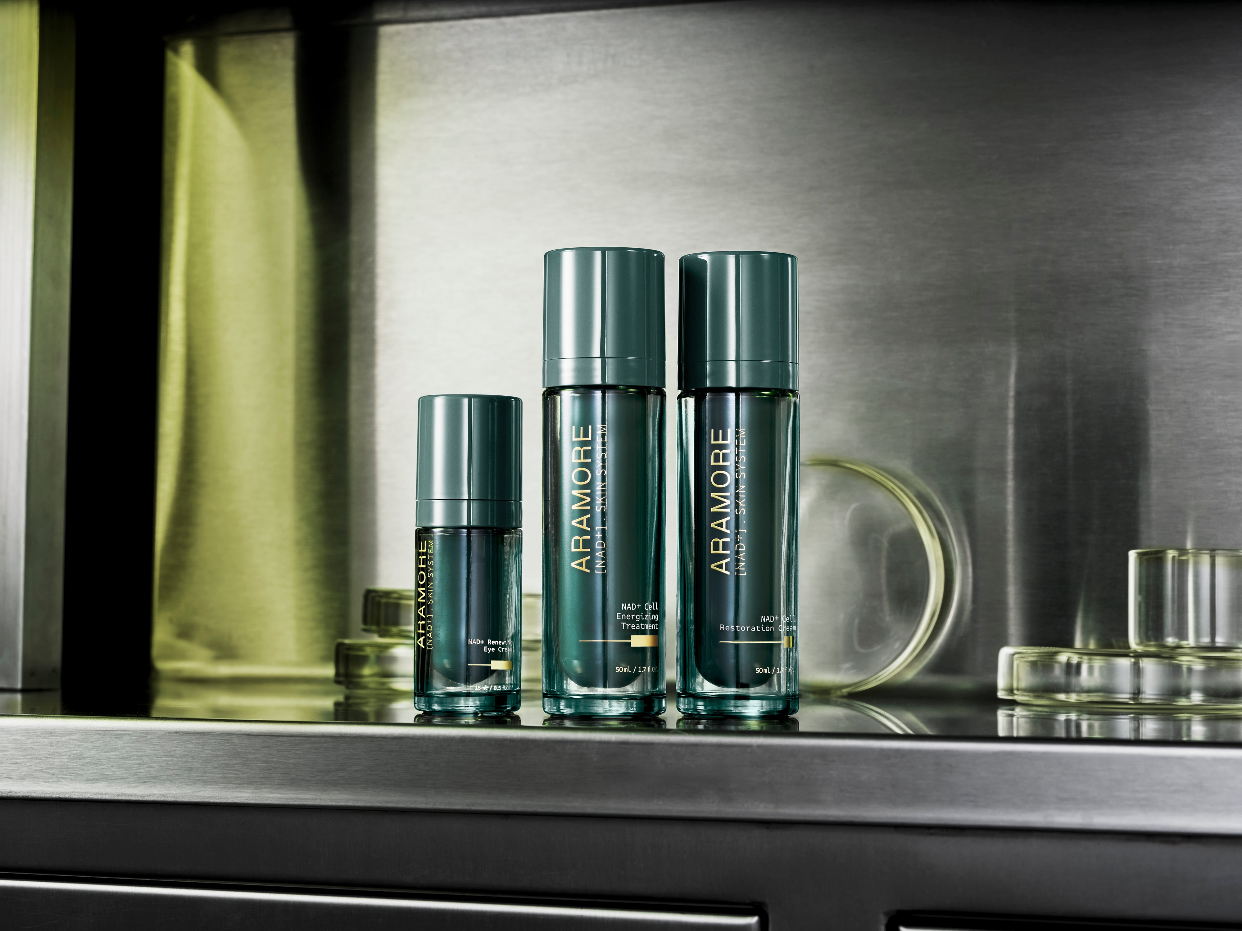 Three Aramore NAD+ Skin System products — the Renewing Eye Cream, Cell Energizing Treatment, and Cell Restoration Cream — displayed on a stainless steel shelf alongside glass beakers.